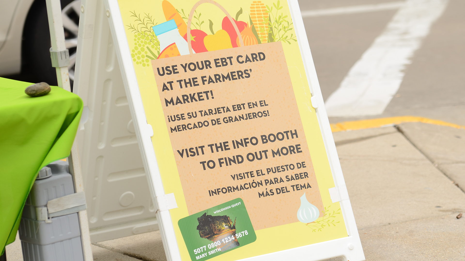 A sandwich board sign on a sidewalk shows an illustration of a paper grocery bag filled with food items, with the words "Use Your EBT Cart at the Farmers' Market" and "¡Use su tarjeta EBT en el mercado de granjeros!"