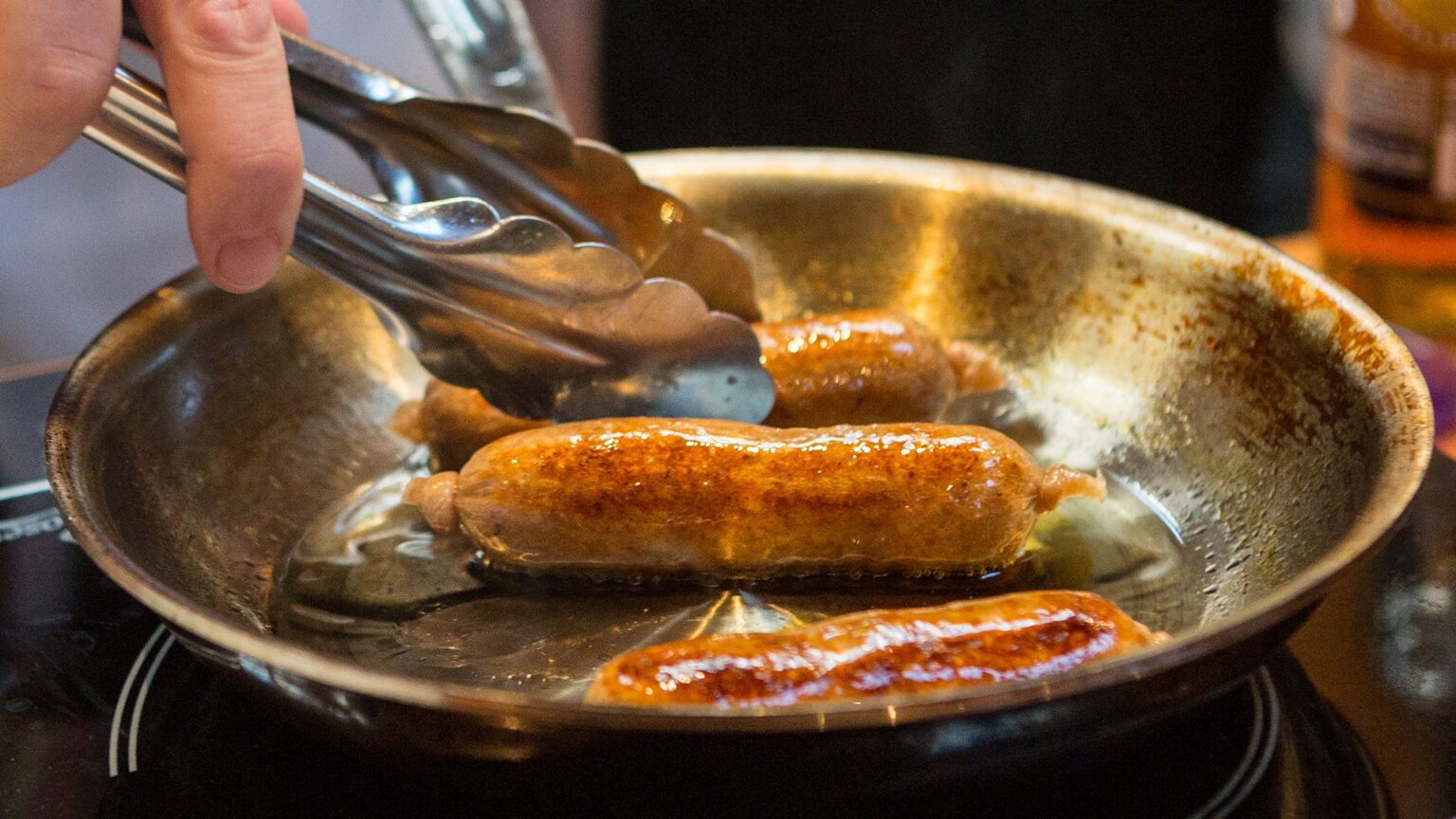 A hand holding a pair of metal tongs turns one of three cultivated meat pork sausages cooking in a metal frying pan.