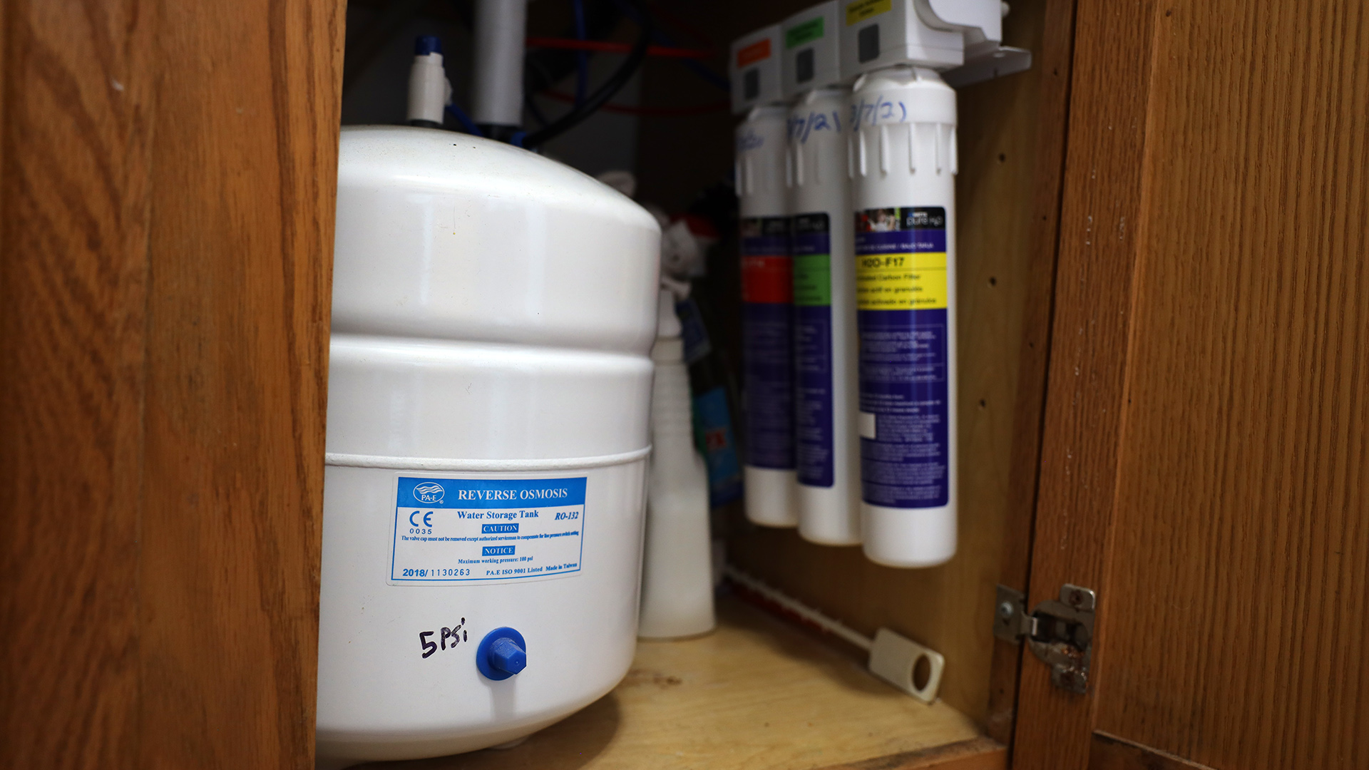 A multi-gallon water container and three adjacent cylinders are installed in a cabinet space under a sink.