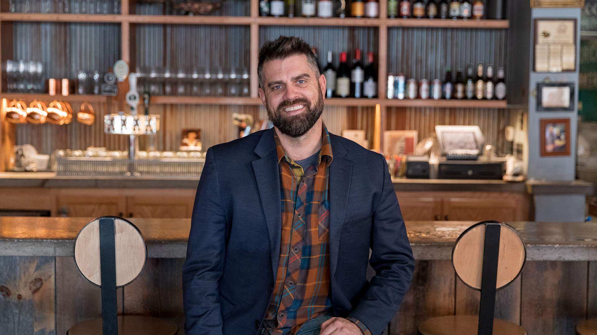 Wisconsin Foodie host Luke Zahm wears a button-up shirt and blazer while seated at a well-lit bar.