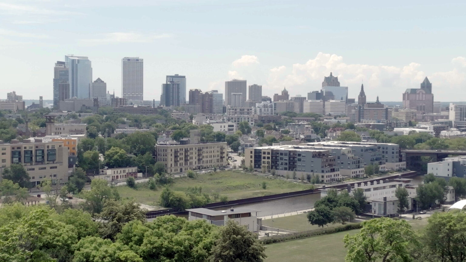 An aerial photo shows a river with with multi-story buildings on other side and a skyline of taller buildings in the background.