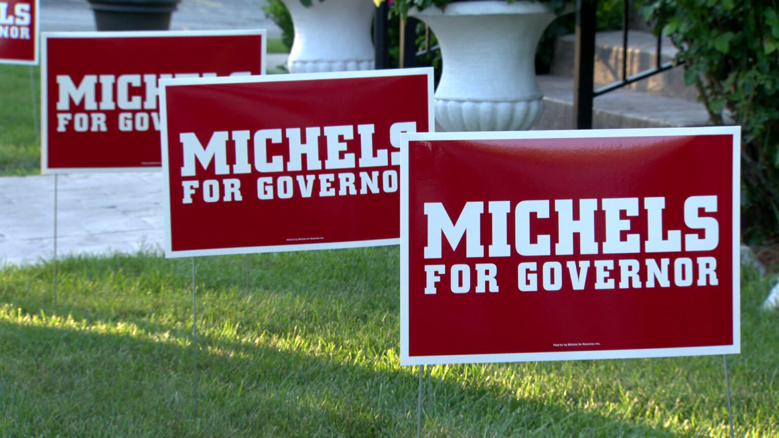 Multiple campaign yard signs reading Michels for Governor are placed in a lawn, with a walkway, stairs and planters in the background.