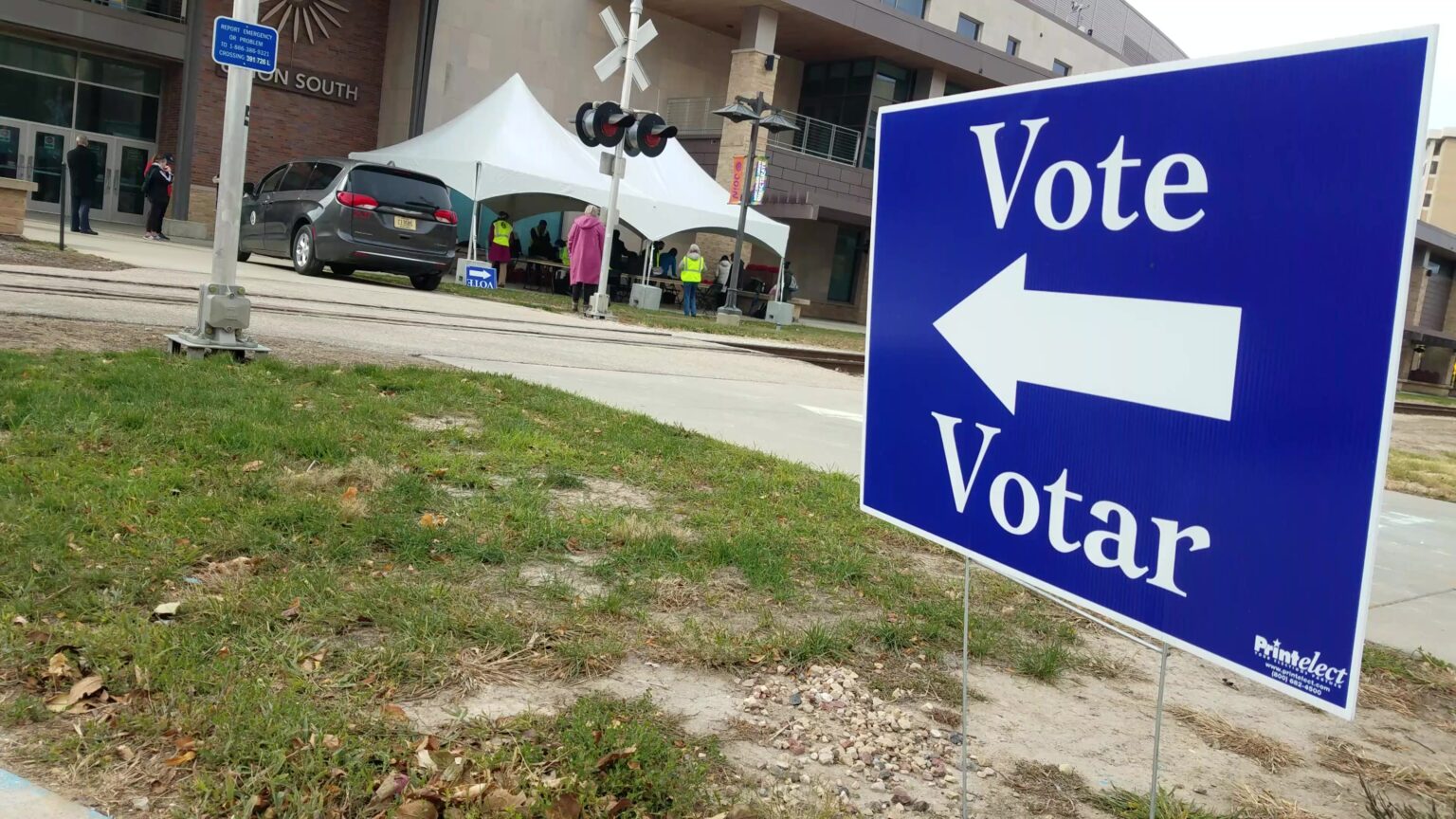 A sign with an arrow and the words Vote and Votar is placed in a patch of lawn across the street from a building with a sign reading Union South and an outdoor canopy with people standing under and around it.