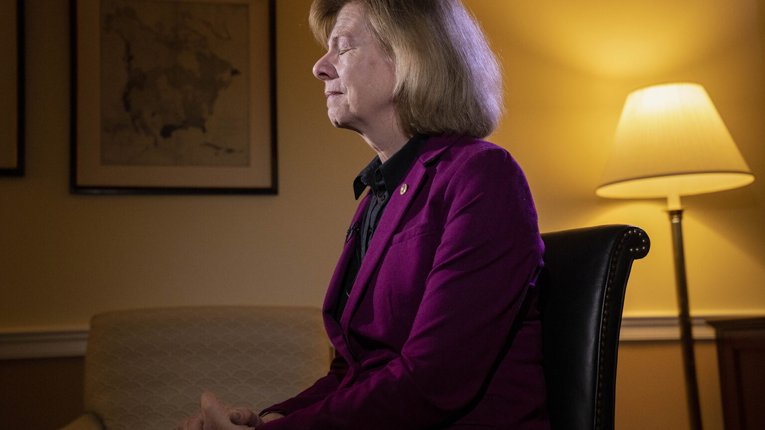 Tammy Baldwin sits in a chair in an office with her eyes closed and hands folded in her lap, with a lit lamp and vintage map of North America in the background.