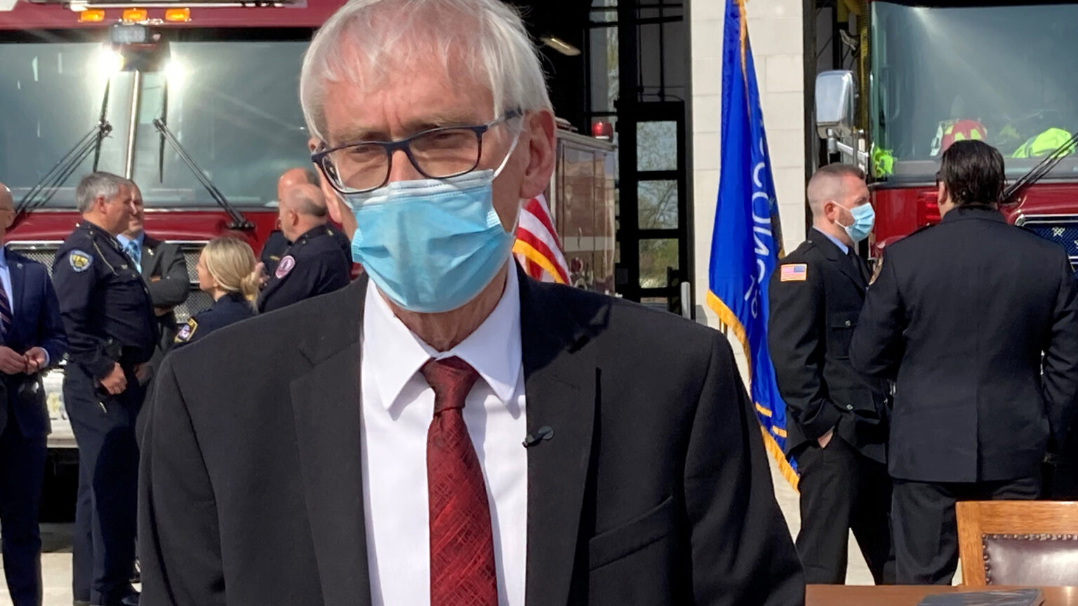 Tony Evers wears a medical face mask while standing in front of a table and chair set outside the garage doors of a fire station, with firefighters standing in the background alongside two parked fire engines and U.S. and Wisconsin flags.