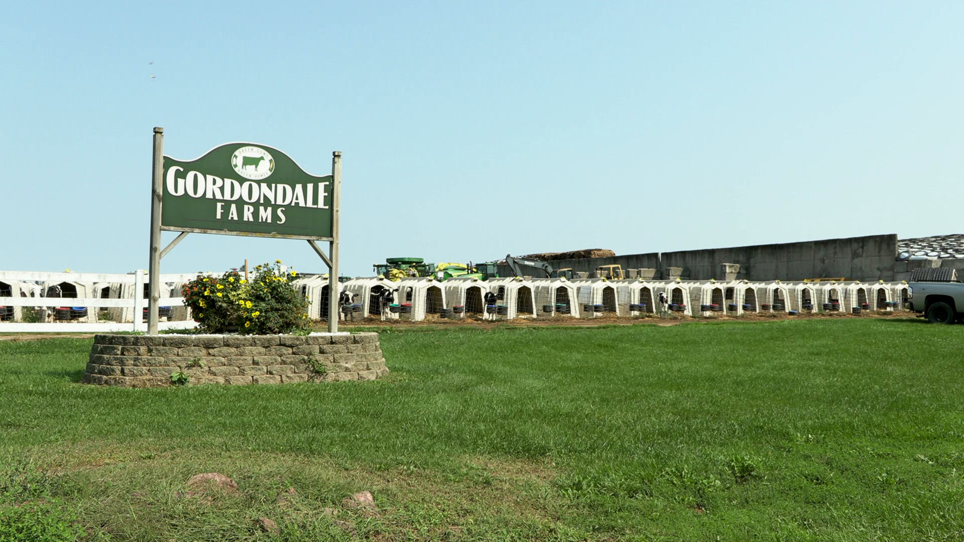 A sign reading Gordondale Farms is mounted in a masonry base and stands in front of a row of calf hutches, with a building and vehicles in the background.
