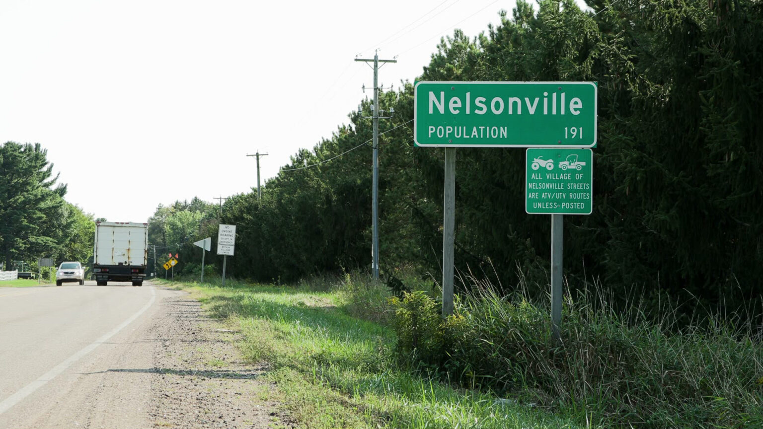 A roadside sign reading Nelsonville Population 191 stands alongside a road next to another sign addressing ATV/UTV traffic, with multiple vehicles, trees and powerlines in the background.