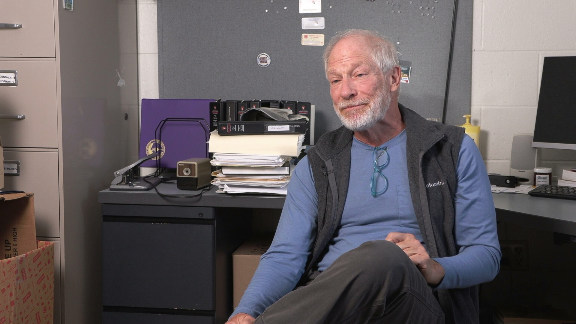 George Kraft sits in an office with a desk, metal filing cabinet, bulletin board and other items in the background.