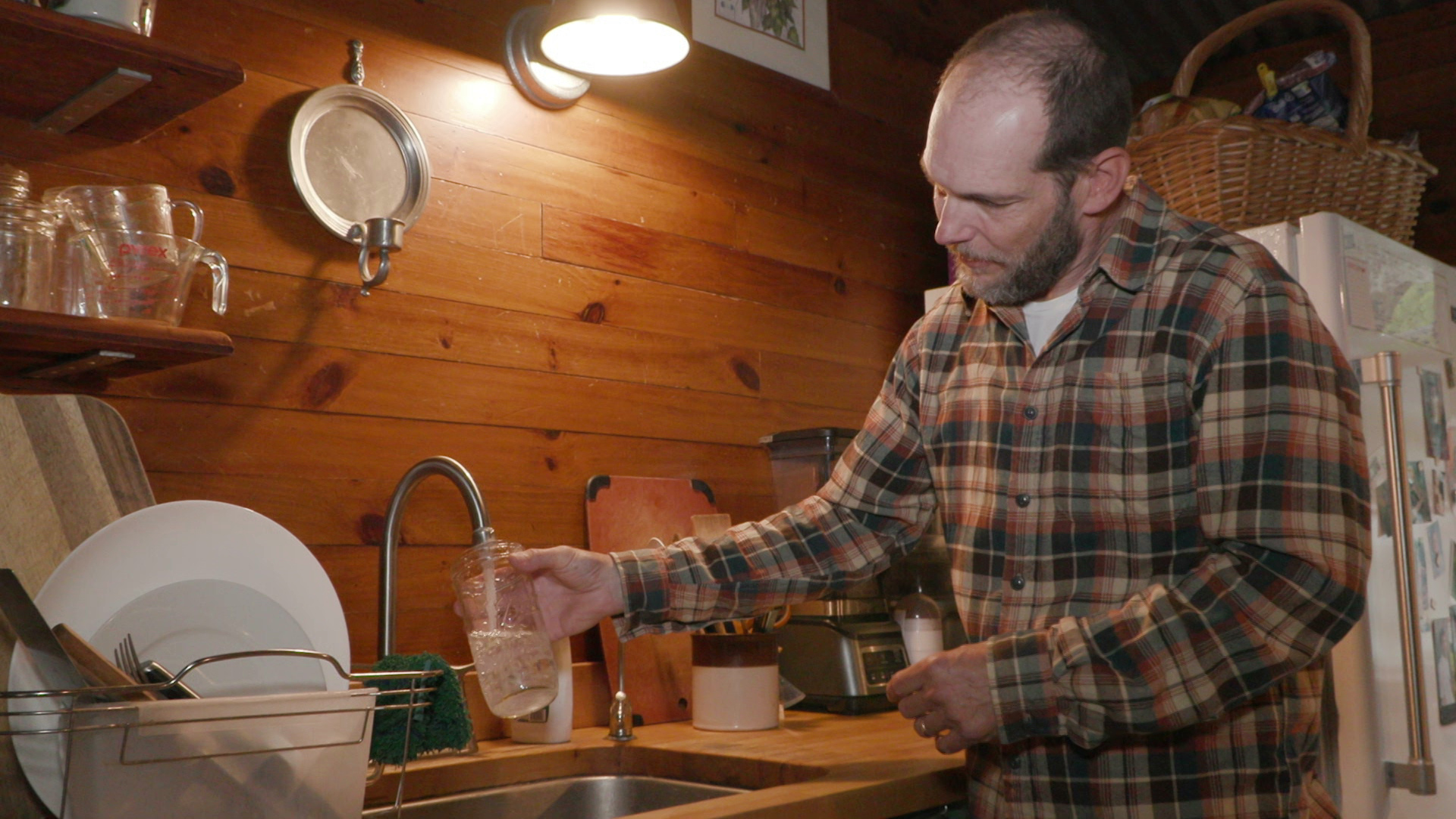 Tarion O'Carroll holds a glass canning jar under a faucet and fills it with water while standing in a kitchen with wood-paneled walls, with shelves and a refrigerator in the background.