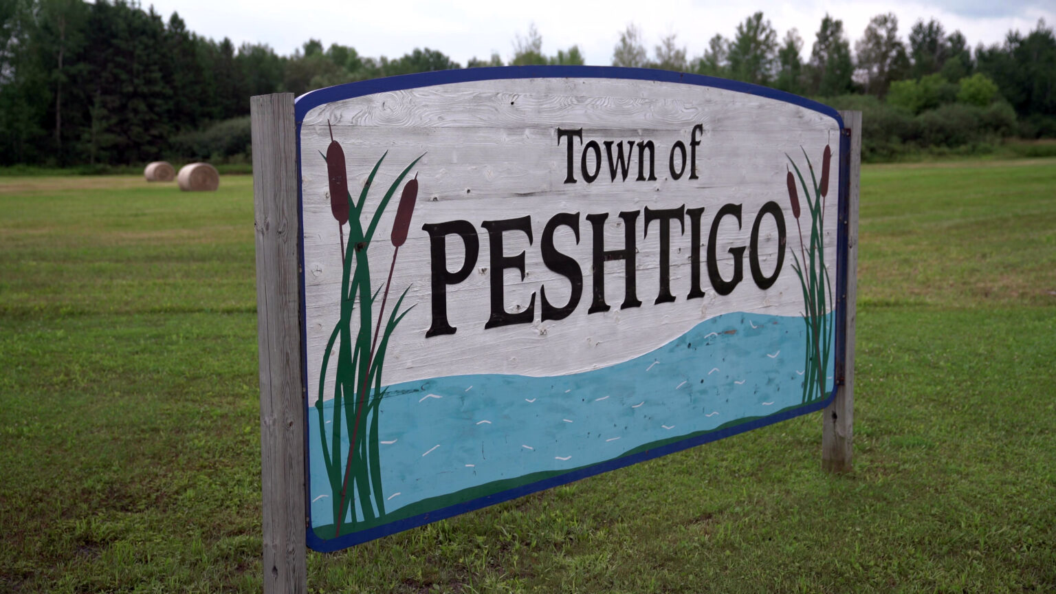 A wood sign with a painting of water and cattail wetland plants with the words Town of Peshtigo stands in a field, with round hay bales and trees in the background.