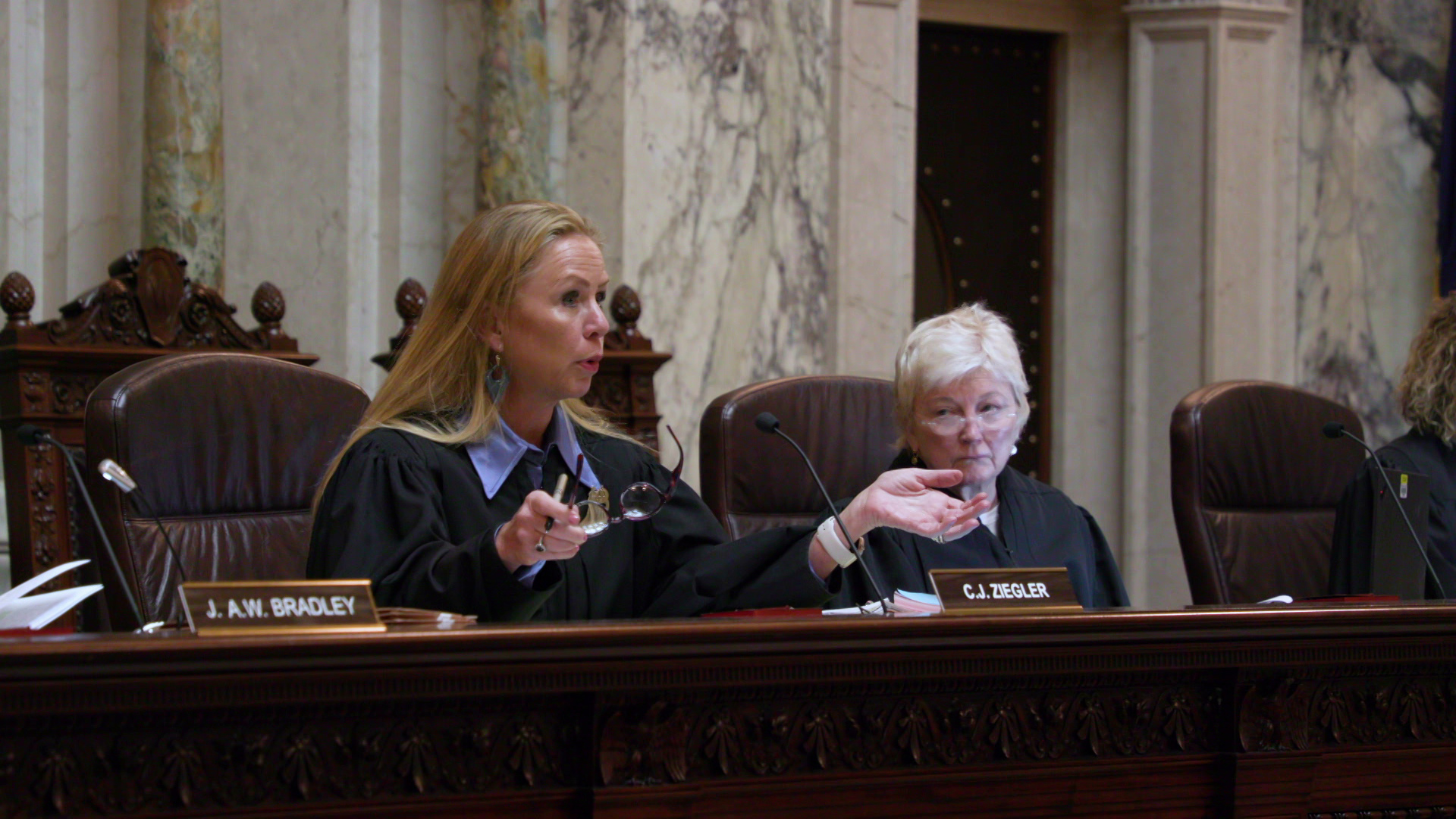Annette Ziegler sits in her chair at the Wisconsin Supreme Court bench and speaks into a microphone while gesturing with both hands, one of which is holding a pen and glasses, with Patience Roggensack listening while seated next to her.
