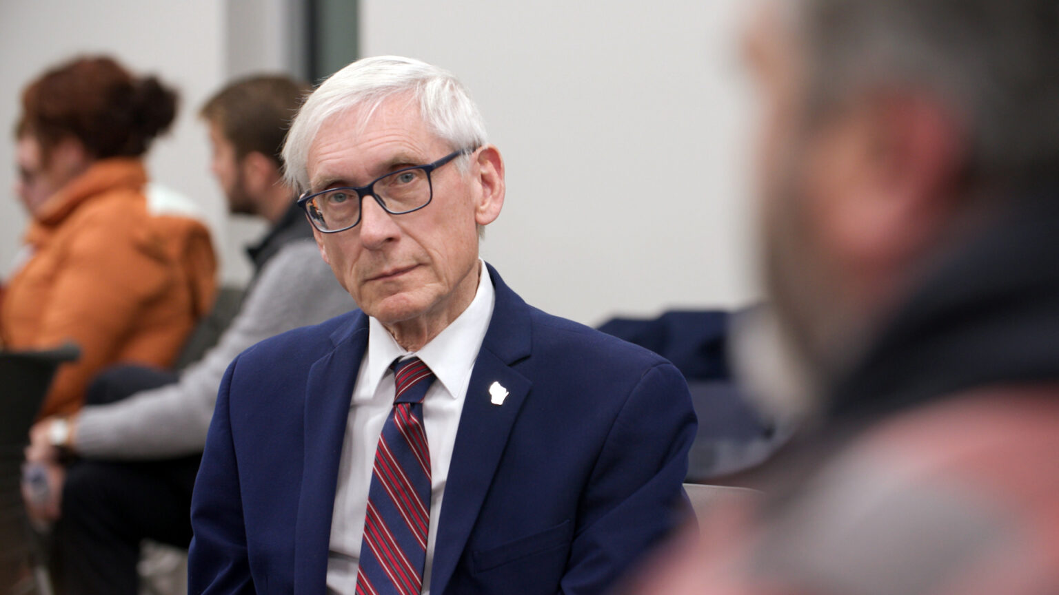 Tony Evers listens to another speaker while seated alongside others, with more people seated in the background.
