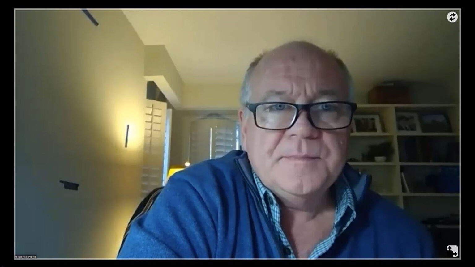 A videoconferencing screen shows Fred Prehn seated in a room and looking into the computer camera, with shelves and window shutters in the background.