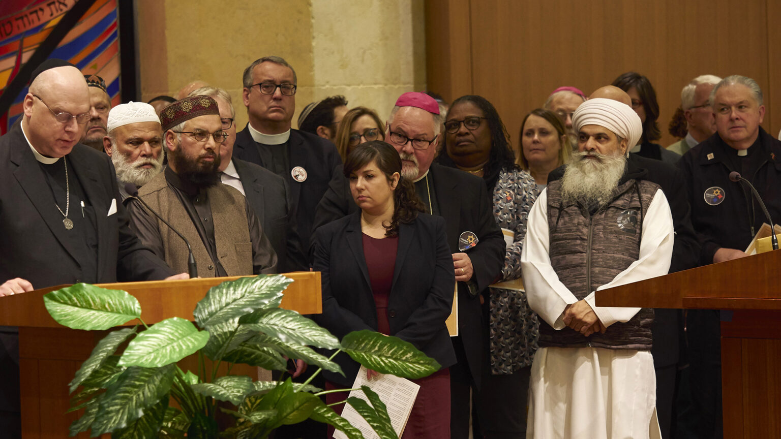More than one dozen people, many clad in religious garments, stand on a stage with two podiums, with a rabbi standing behind one and speaking into a microphone.