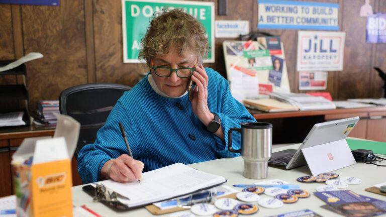 Mary Lynne Donohue sits at a desk covered with political buttons and fliers, a laptop computer, thermal coffee mug and other items, while holding a mobile phone to her left ear and writing on a piece of paper on a clipboard with her right hand, with office items on desks and political posters on walls in the background.