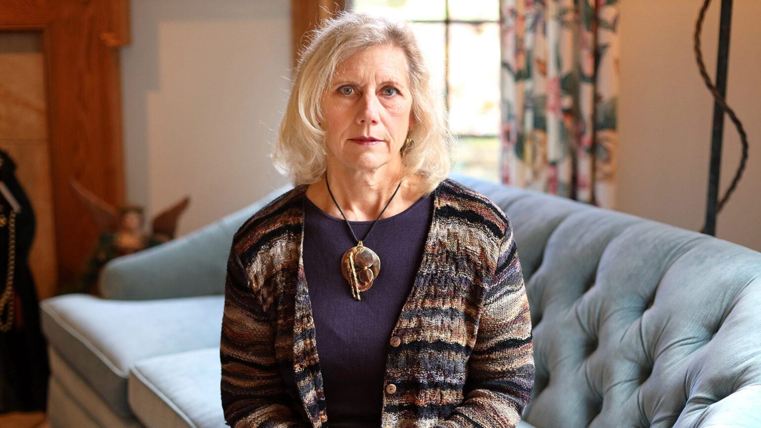 Kathy Hartke poses for a portrait while seated on a couch, with a window with a half-open curtain in the background.