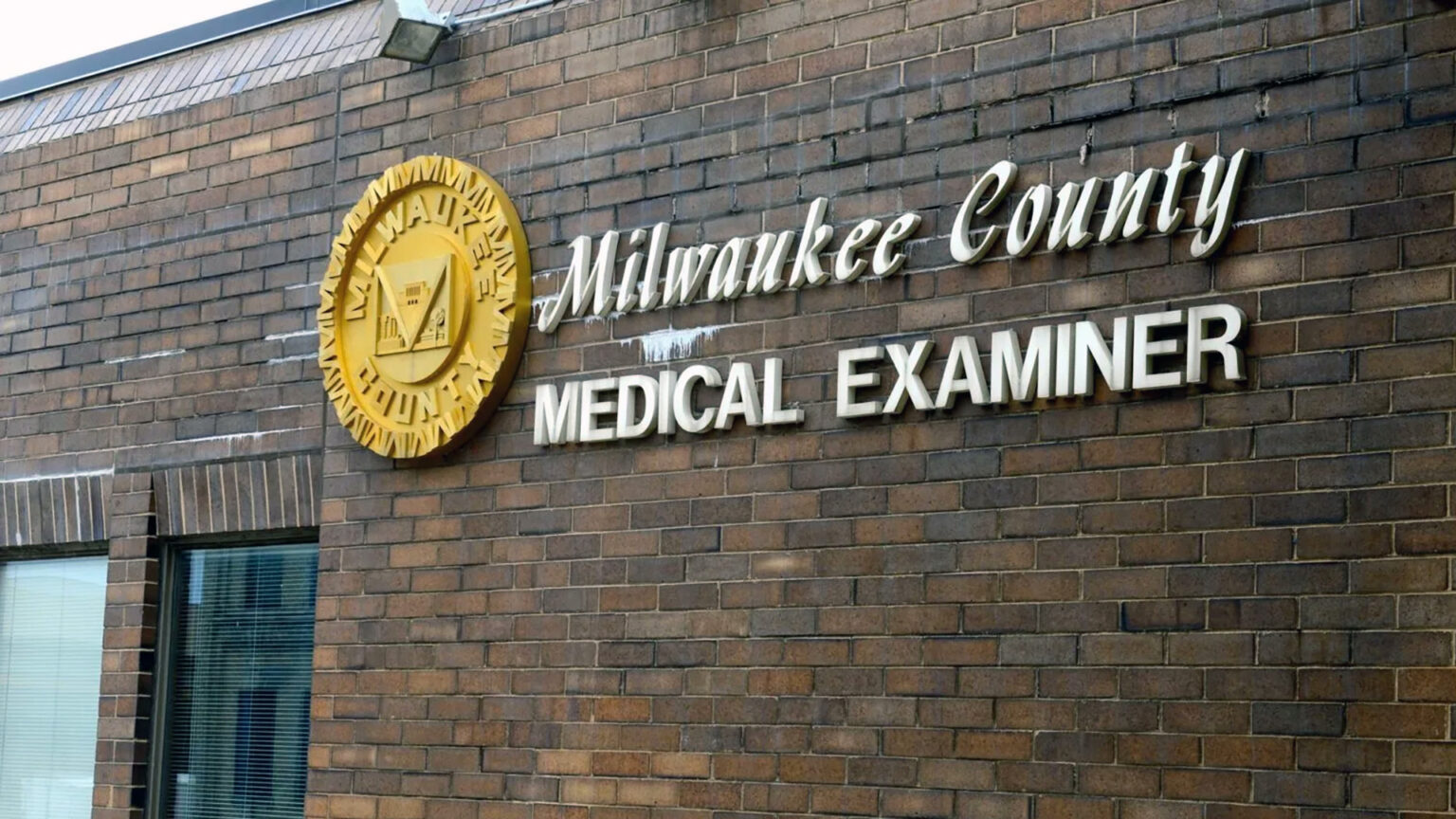 A sign with a relief seal of Milwaukee County and letters spelling Milwaukee County Medical Examiner sit on the side of a brick wall of a building.