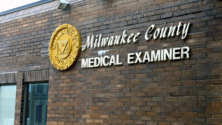 A sign with a relief seal of Milwaukee County and letters spelling Milwaukee County Medical Examiner sit on the side of a brick wall of a building.