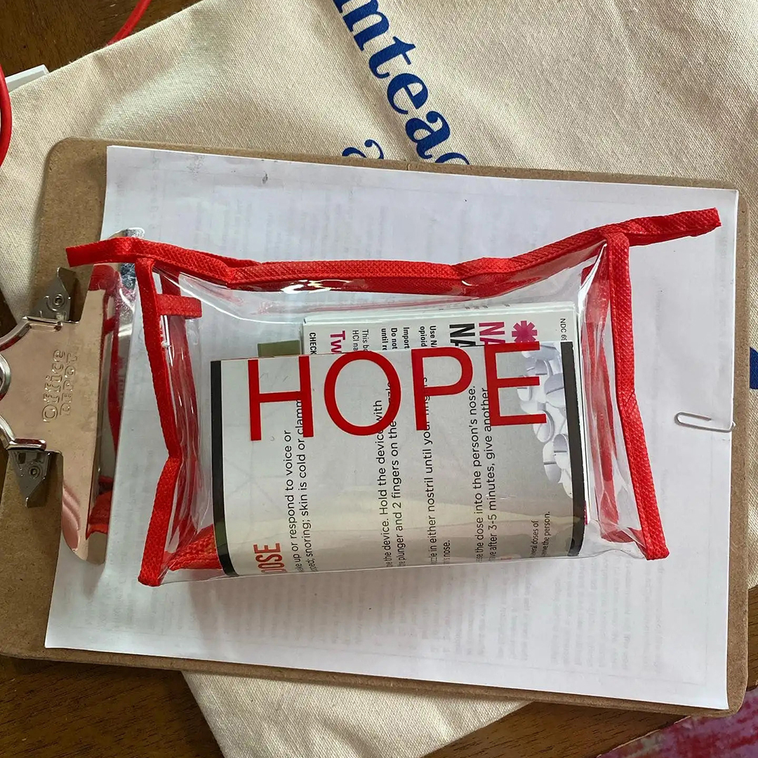 A transparent plastic back with a zippered lining is filled with multiple items and sits on top of a clipboard with multiple sheets of paper and a canvas tote bag.