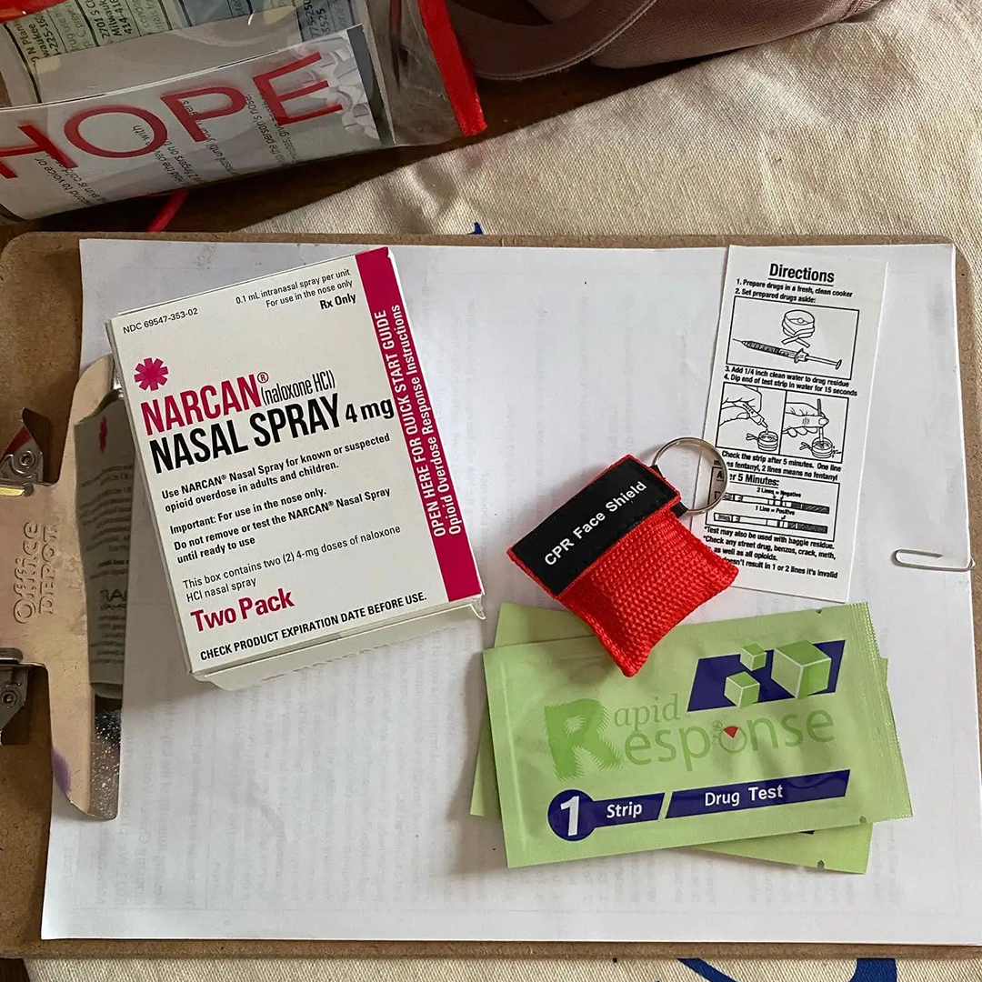 A box of Narcan nasal spray, a CPR face shield, two "Rapid Response" fentanyl testing strip packages and a piece of paper labeled "Directions" sit on top of a clipboard with multiple sheets of paper.