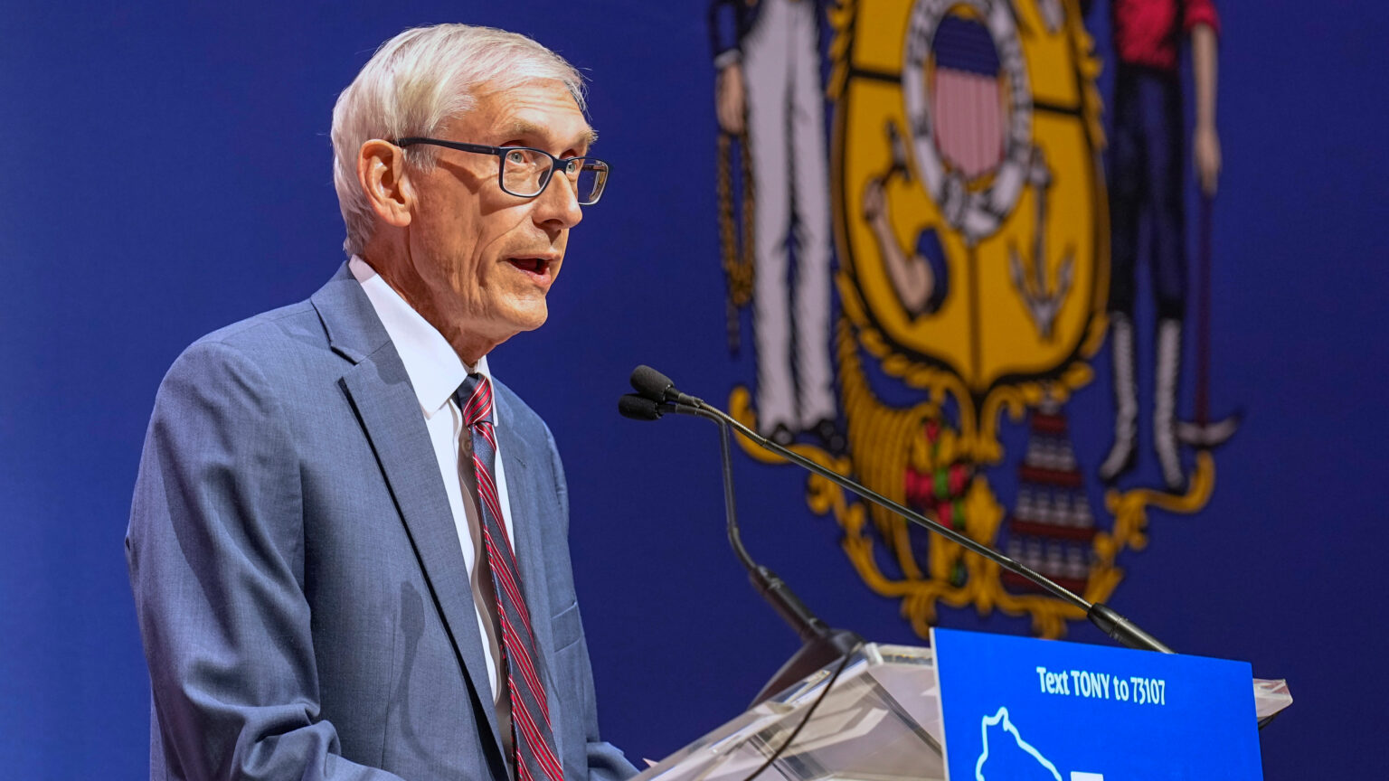 Tony Evers talks into a microphone on a podium in front of a Wisconsin state seal.