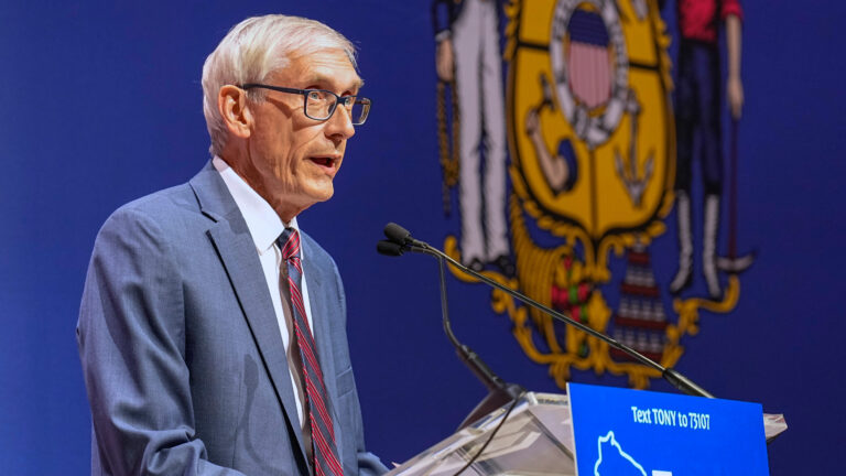 Tony Evers talks into a microphone on a podium in front of a Wisconsin state seal.