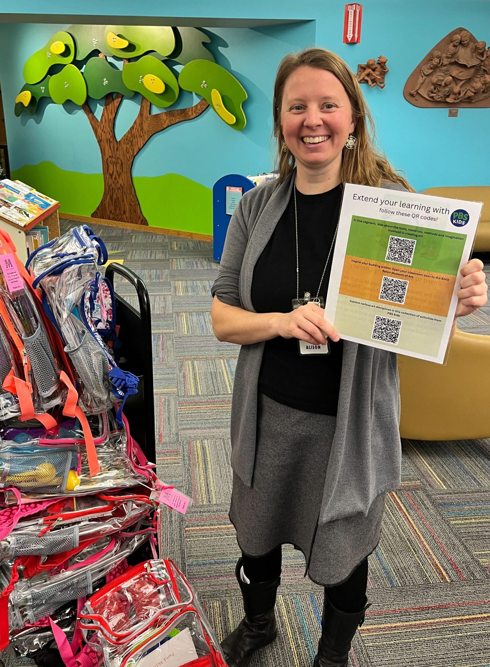 A librarian stands next to a shelf of backpacks and holds up a sheet of paper.