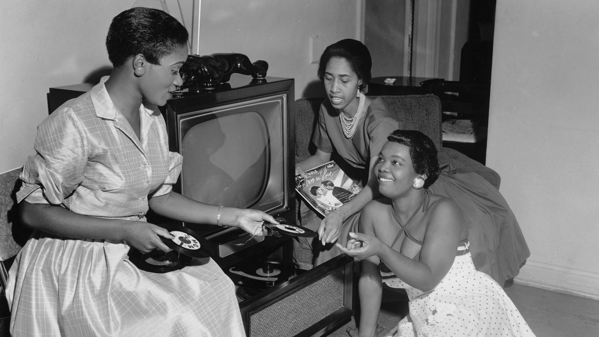 A group of women discuss vinyl records in a black and white picture taken in the 1940s or 1950s.