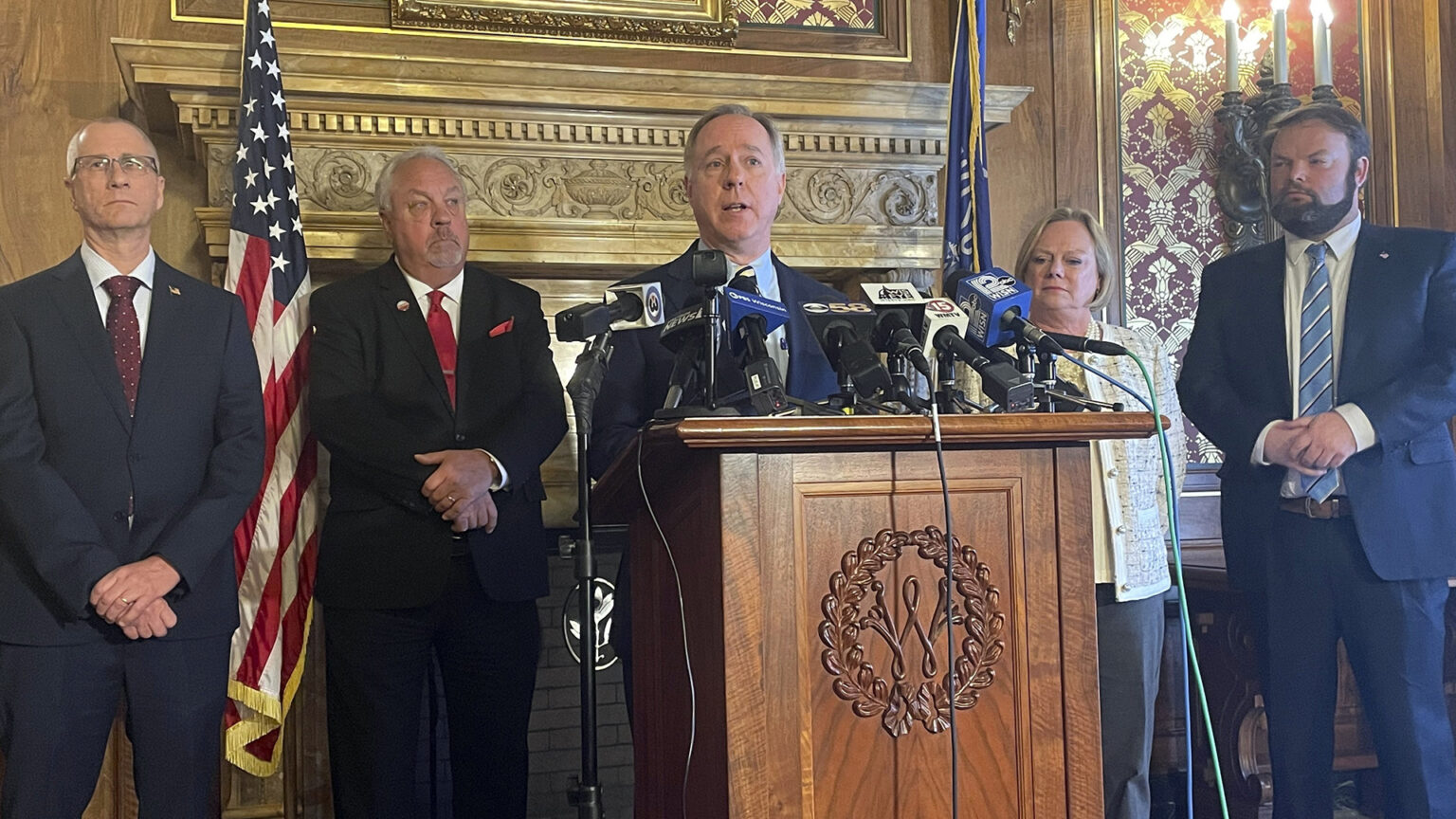 Robin Vos stands behind a wood podium with multiple microphones mounted on it with other Assembly members standing on either side of him in a room with wood-paneled walls, sconce-style lighting fixtures, a painting and the U.S. and Wisconsin flags.