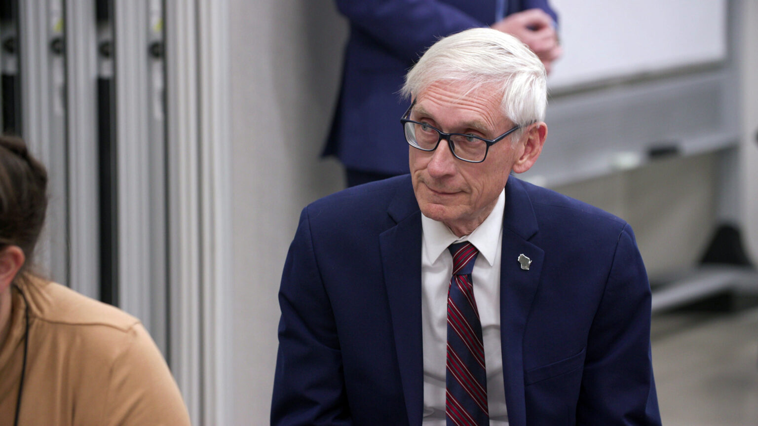 Tony Evers sits and listens in a room with other people siting and standing in the foreground and background.