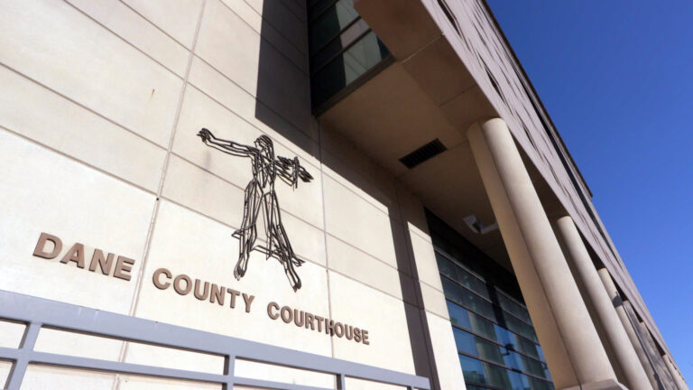 A sign reading Dane County Courthouse and a stylized flat, wire sculpture of Lady Justice are mounted on the masonry wall of a Brutalist-style building that also features pillars and multi-story windows.