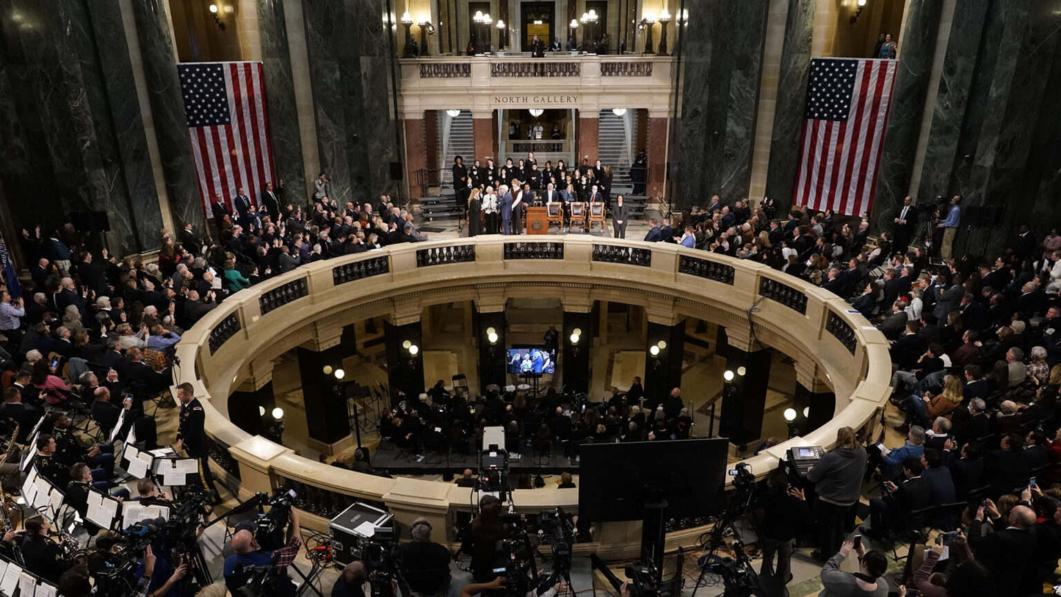 Tony Evers stands and delivers his oath of office while standing next to a railing in the Wisconsin Capitol Rotunda, which is decorated with two large U.S. flags and with audience members, musicians and journalists seated and standing around the circle and on the ground floor level below.