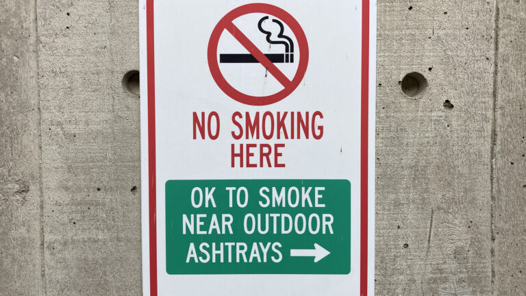 A metal sign attached to a concrete wall shows a circle-slash no symbol over a lit cigarette illustration with the words No Smoking Here and OK to Smoke Near Outdoor Ashtrays with an arrow pointing to the right.