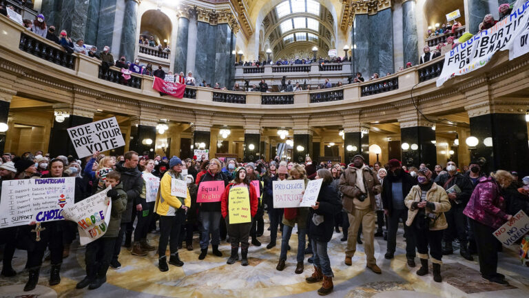 Numerous people, including those holding a variety of homemade protest signs, stand on multiple levels of a rotunda with marble pillars, walls, balustrades and lit by wall-mounted lamps and skylights.