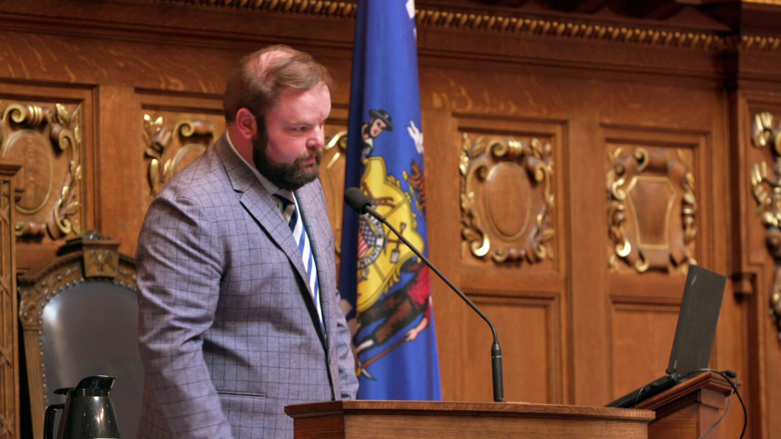 Tyler August stands in front of a dais and speaks into a microphone while facing an open laptop computer, with a Wisconsin flag and wood-paneled wall with bas-reliefs of stylized shields in the background.