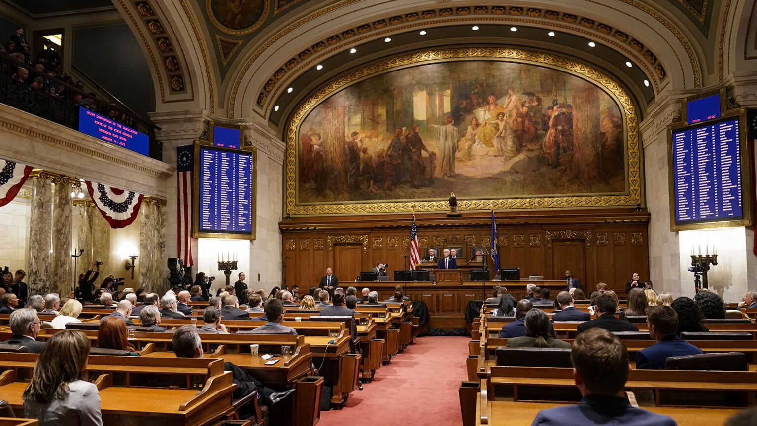 Tony Evers speaks while standing at a podium at the far end of a room as legislators are seated behind him and at rows of desks facing him, in a room with marble pillars and vaulted-arch supports, and a large fresco-style painting and taxidermy bald eagle on the wall above the dais.