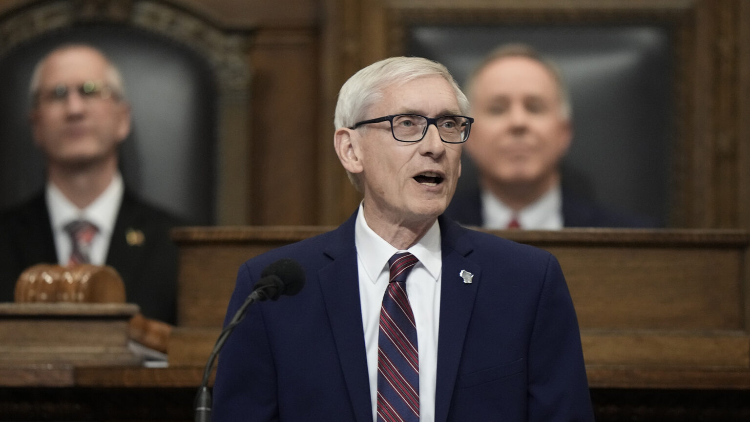 Tony Evers stands and speaks into a microphone with two other people seated behind him on a dais.