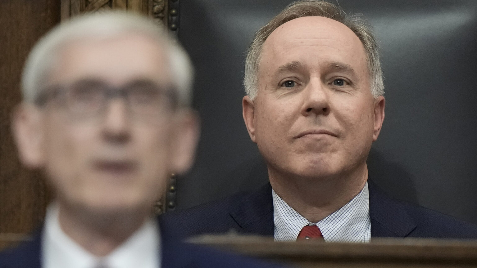 Robin Vos is seated in a high-backed chair behind Tony Evers, who is speaking (and out of focus) in the foreground.