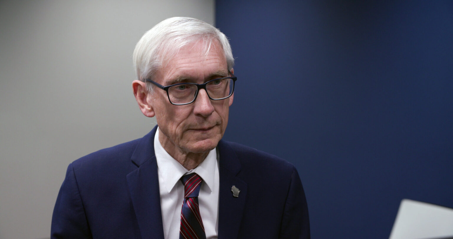 Tony Evers stands in a room and responds to questions from a reporter.