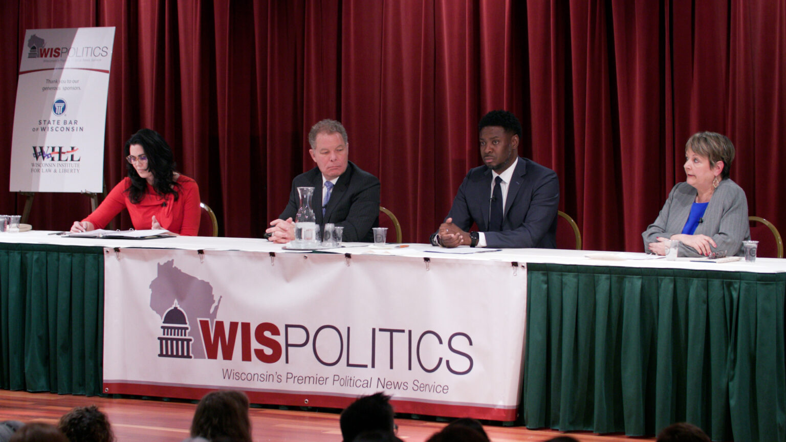 Jennifer Dorow, Daniel Kelly, Everett Mitchell and Janet Protasiewicz sit at a table ringed with stage drapery and a banner with a wordmark for WisPolitics, with a sponsor sign for WisPolitics, the State Bar of Wisconsin and the Wisconsin Institute for Law and Liberty in the background.