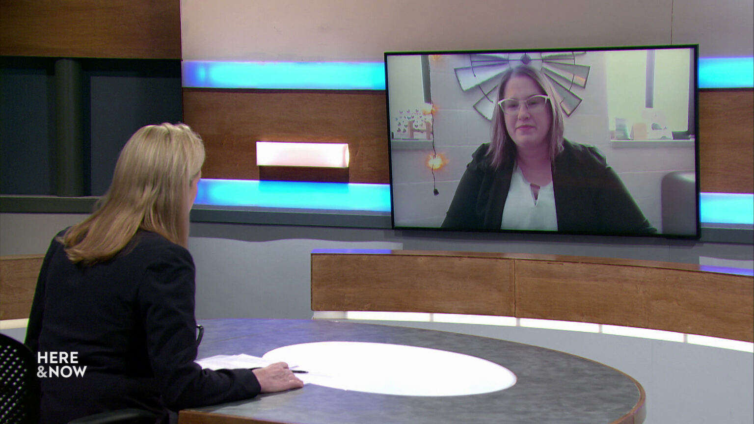 Frederica Freyberg sits at a desk on the Here & Now set and faces a video monitor showing an image of Cindy Bourget.