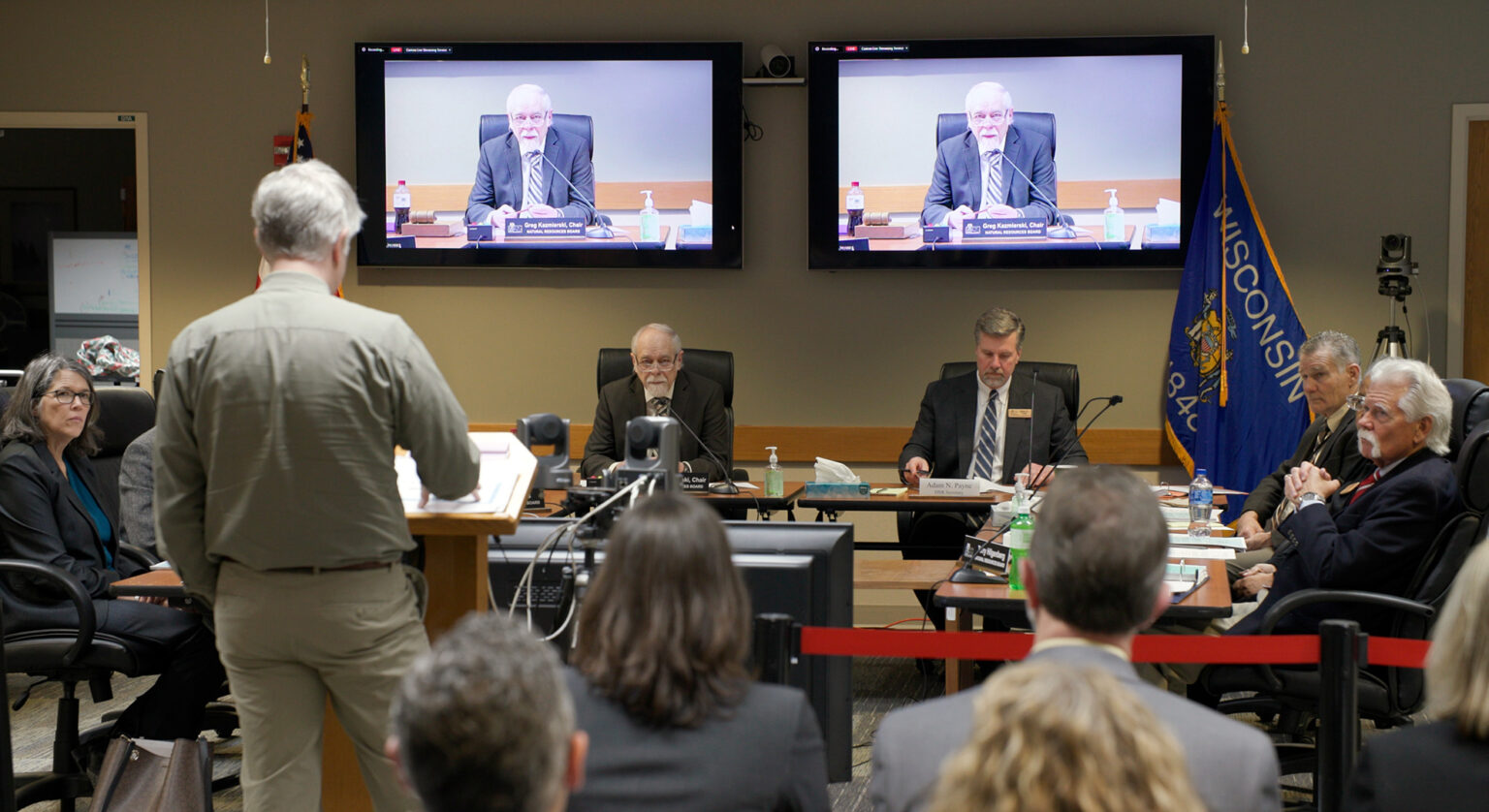 A person standing at a podium with their back turned to the camera faces a group of people seated on three tables arranged in an open square, with a Wisconsin flag on the opposing wall along with two television monitors showing one of the seated individuals, with other seated people in the foreground facing the discussion.