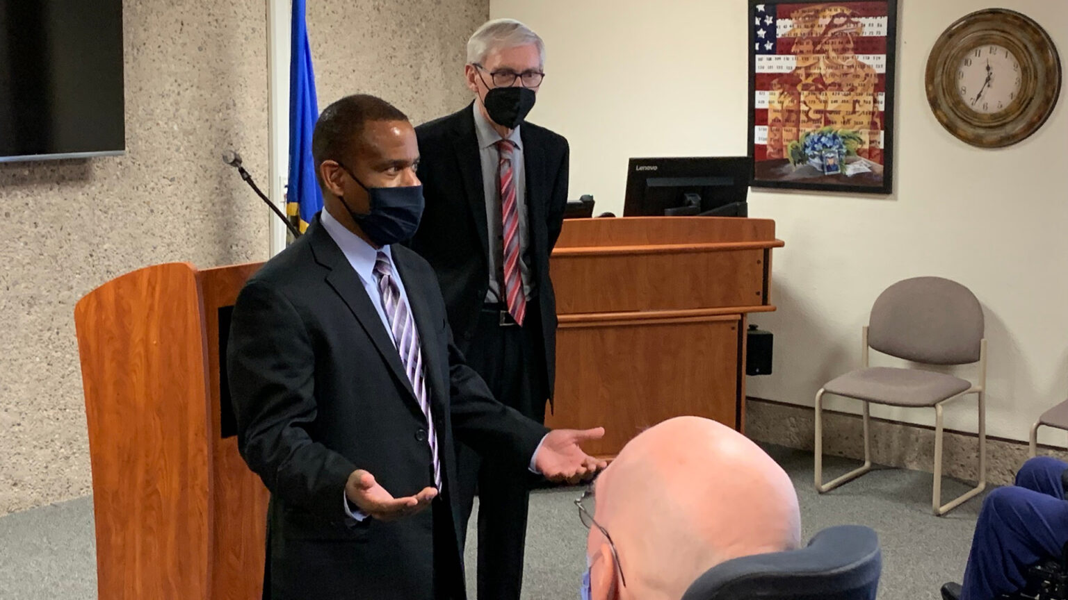 James Bond speaks in front of a podium to veterans in a meeting room while gesturing with both hands and wearing a cloth face mask, with Tony Evers standing in the background and wearing a face mask, with a video monitor, Wisconsin flag, desk, chairs, clock and veterans-themed poster in the background.
