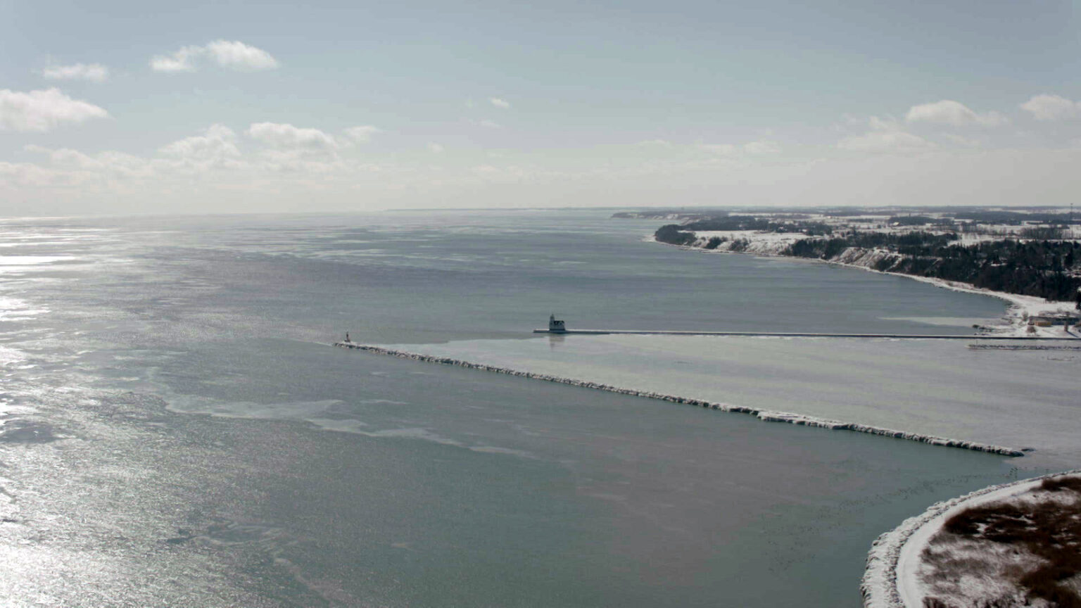 An aerial photo shows two breakwaters extending from a shoreline into a frozen body of water, with sunlight reflecting off the surface of the ice and snow covering the ground on the land.