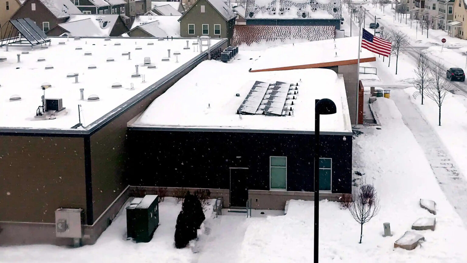 Solar panels are situated on the flat roof of a section of a multi-segment building, with a U.S. flag, houses and a street in the background, all covered with a layer of snow.