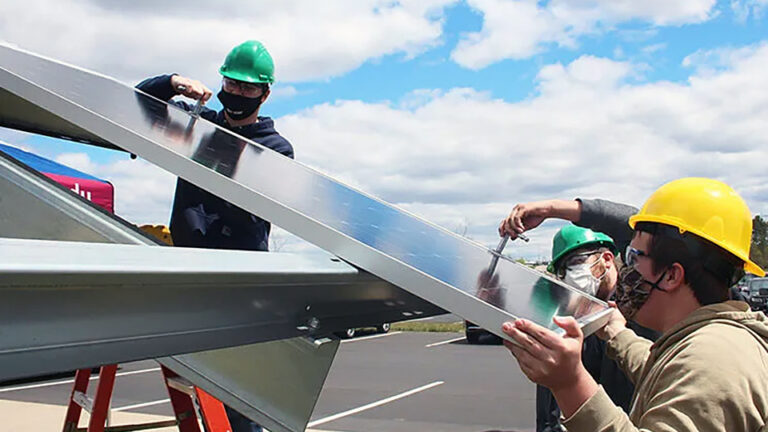 Three people hold a solar panel on a mounting and use tools to adjust its placement while standing next to a parking lot.