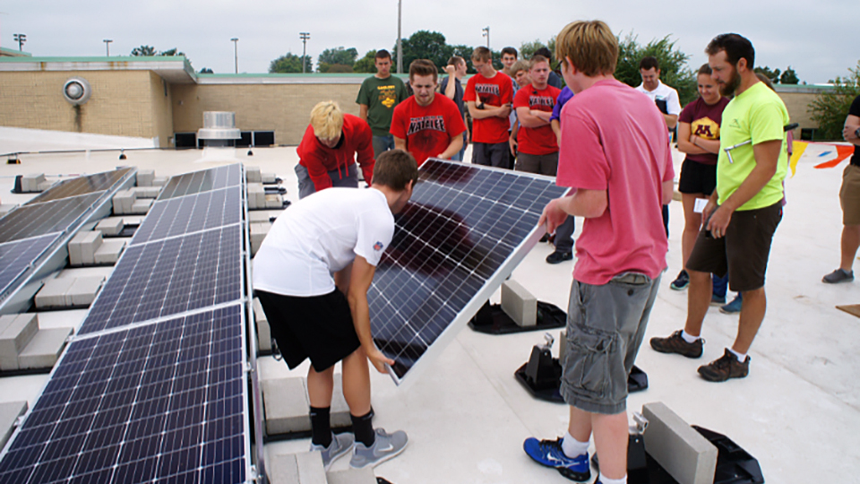 Four people holding the corners of a solar panel lower it on to a mounting alongside two adjacent rows of solar panels on a rooftop, as a group of people stand in the background and watch, with another flat roof, light polls and trees in the background.