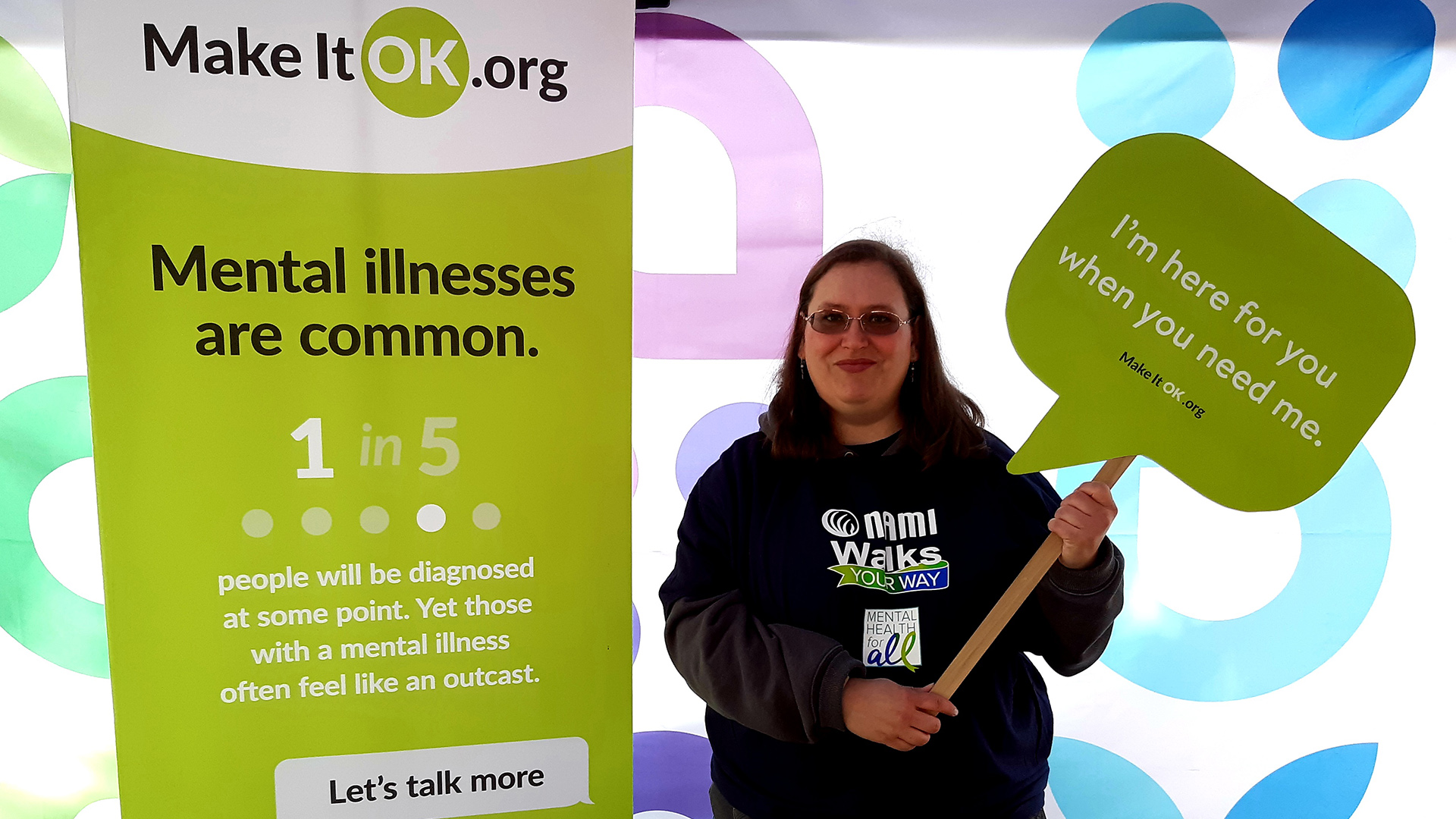 Chrissy Barnard holds a sign in the shape of a word bubble reading "I'm here for you when you need me." and the URL makeitok.org while standing next to a banner sign with the same URL, the words "Mental illnesses are common." in large type and other details about the issue.