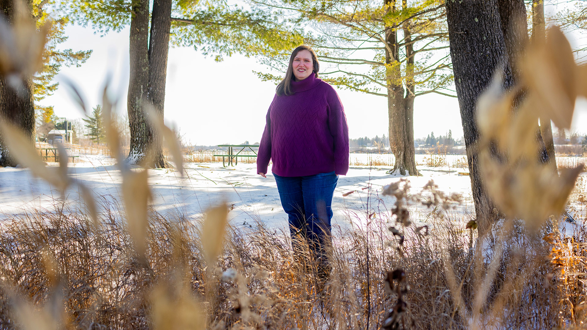 Chrissy Barnard stands outdoors in front of a snow-covered field among multiple trees, with a picnic table in the background and more trees on the horizon.