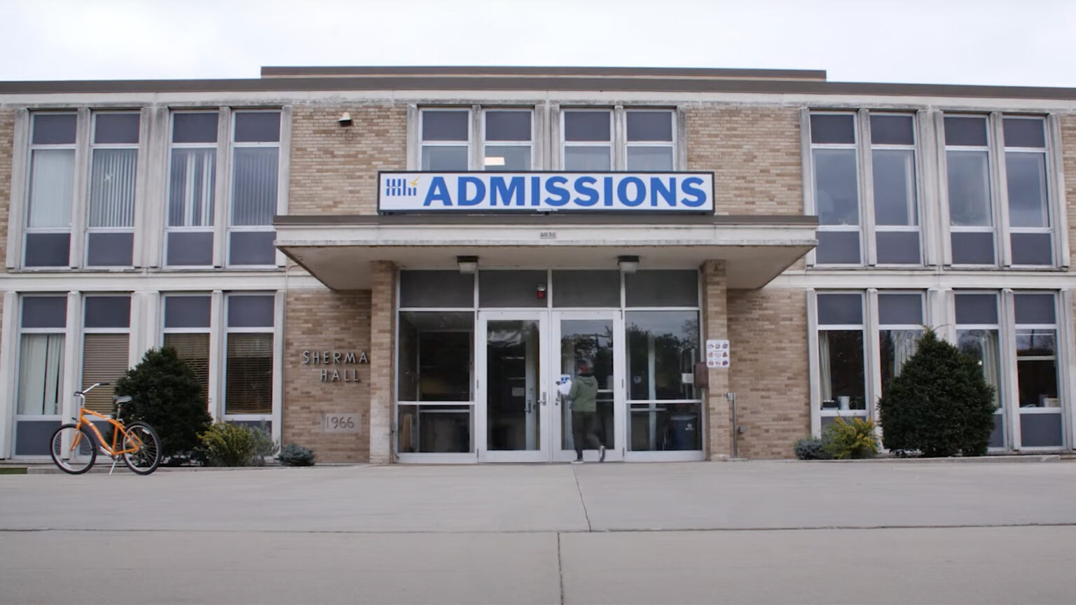 A sign reading Admissions sits atop the awning to an entrance of a two-story brick building with tall glass-and-metal panel windows, with a person walking toward the doors and a bicycle parked in front of bushes to the side.